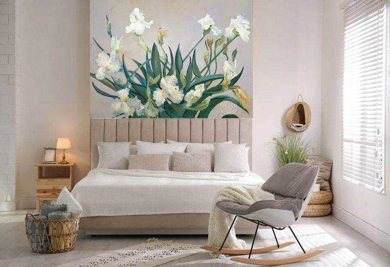 A styled bedroom interior featuring a large wall mural of white irises on a beige background, behind a bed with white linens and a gray upholstered rocking chair in the foreground.
