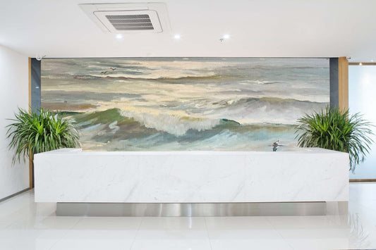 A large ocean wave painting displayed on a wall behind a white reception desk, flanked by two green potted plants in a bright modern lobby.